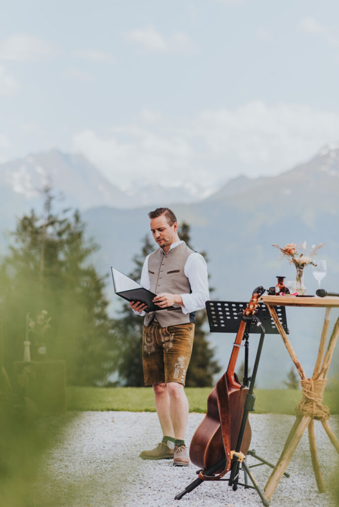 Ein Mann in traditioneller Kleidung liest im Freien in Süddeutschland aus einem Notizbuch, während in der Nähe Berge und Instrumente von Hochzeitsmusikern zu sehen sind.
