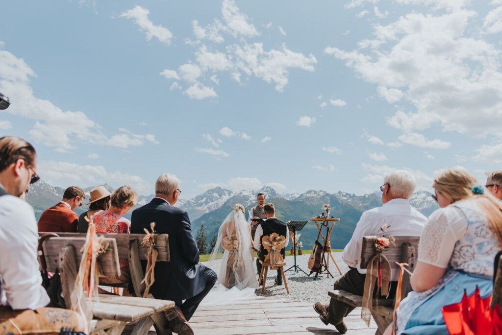 Trauung im Freien mit sitzenden Gästen, Braut und Bräutigam mit Blick auf den Trauredner, im Hintergrund die Berge Südtirols.