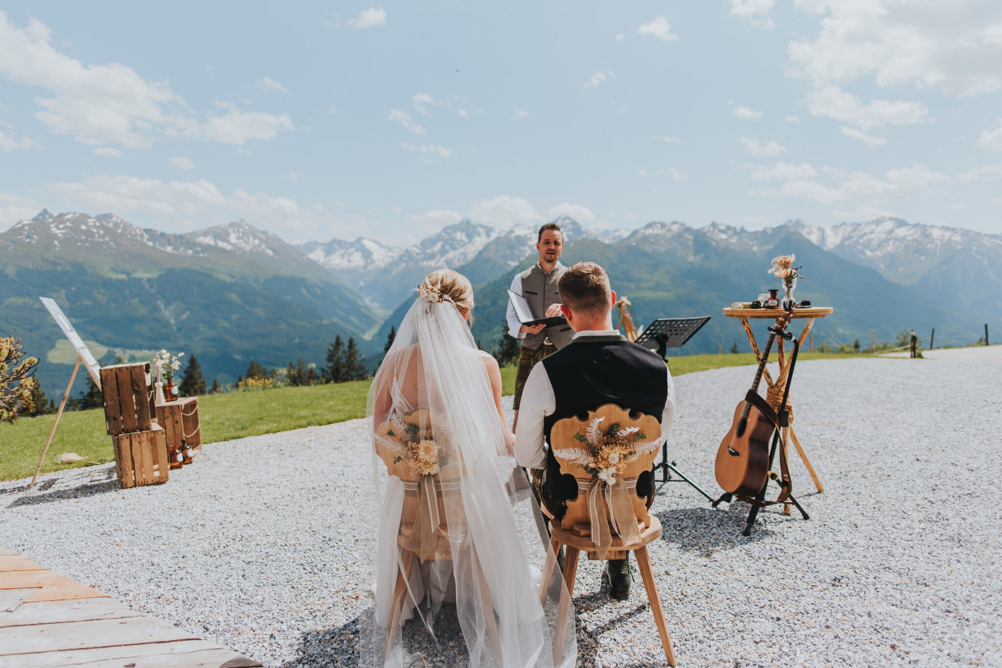 Braut und Bräutigam sitzen im Freien bei einer Hochzeitszeremonie in den Bergen in Südtirol, während ein Geistlicher und ein Hochzeitsmusiker in der Nähe auftreten.