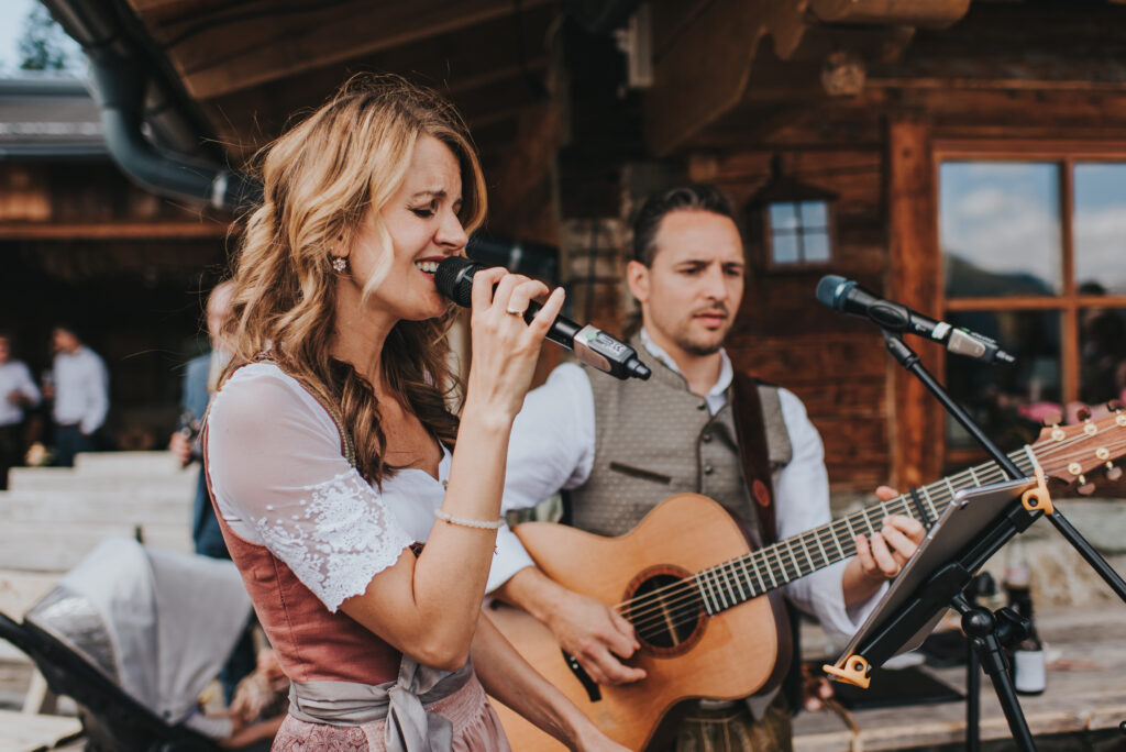 Eine Frau singt in ein Mikrofon, während ein Mann in traditioneller Kleidung im Freien Gitarre spielt und so eine lebhafte Atmosphäre schafft, die typisch für Feste in Südtirol und Süddeutschland ist.