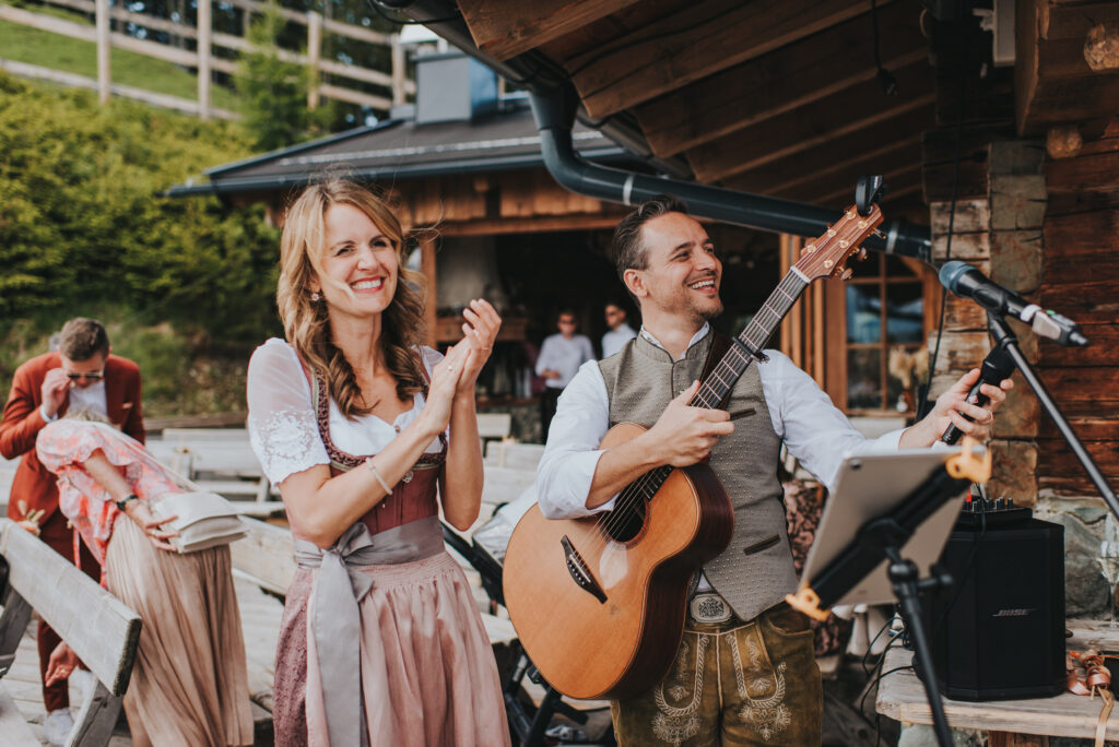 Eine Frau klatscht und ein Mann mit einer Gitarre lächelt im Freien, beide tragen Südtiroler Trachten.