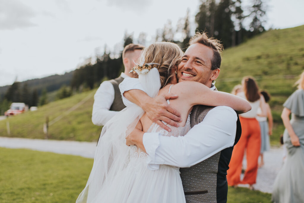 Eine Braut und ein Mann umarmen sich freudig im Freien bei einer Hochzeit in Südtirol, während die Gäste, das üppige Grün und die Hochzeitsmusiker im Hintergrund für eine festliche Atmosphäre sorgen.
