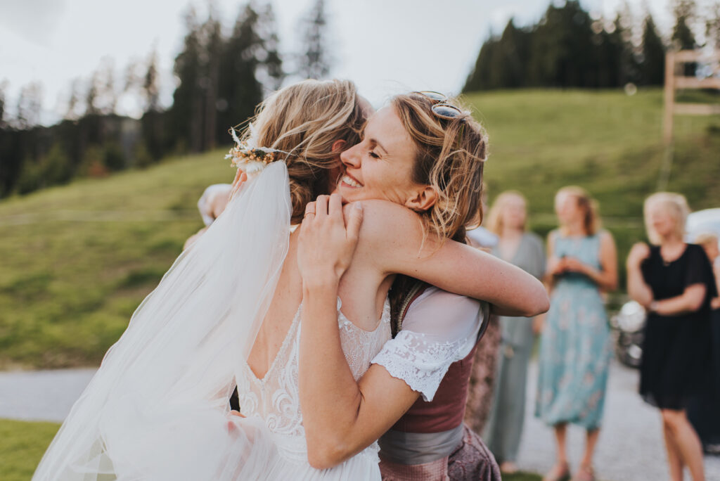 Zwei Frauen umarmen sich im Freien auf einer Hochzeit in Süddeutschland, umgeben von Gästen und üppigem Grün - perfekt, um besondere Momente festzuhalten oder die Klänge eines Hochzeitssängers zu genießen.