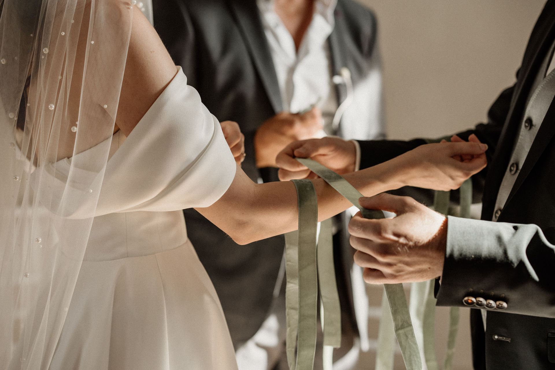 Ein Paar in Hochzeitskleidung nimmt an einer Handfasting-Zeremonie mit salbeigrünen Bändern um die Hände teil, begleitet von einem talentierten Hochzeitssänger und Trauredner, die unvergessliche Erinnerungen in Südtirol schaffen.