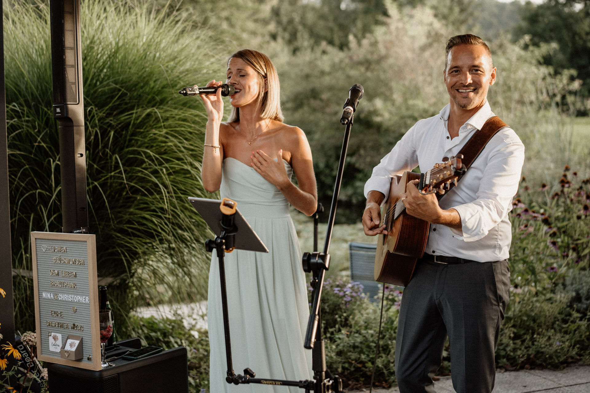 Eine Hochzeitssängerin singt in ein Mikrofon, während ein Mann neben ihr Gitarre spielt und als Hochzeitsmusiker eine zauberhafte Atmosphäre in einem schönen Südtiroler Garten schafft.