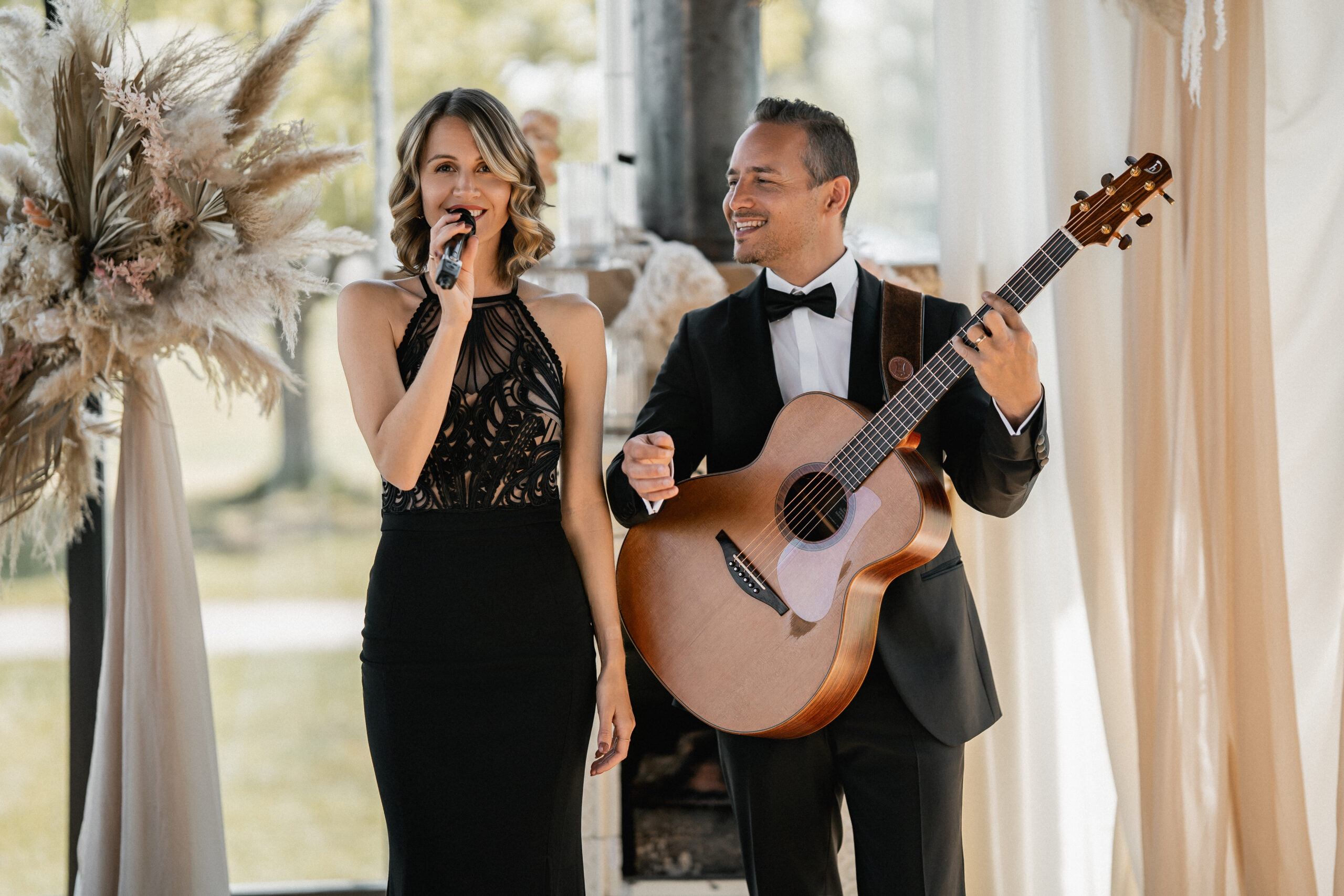 Eine Hochzeitssängerin singt in ein Mikrofon, während ein Mann im Smoking bei einer Indoor-Veranstaltung in Süddeutschland neben ihr Gitarre spielt.