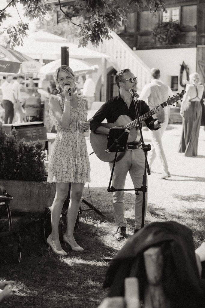 Eine Frau singt und ein Mann spielt Gitarre bei einer Freiluftveranstaltung in Südtirol und unterhält die Gäste als talentierter Hochzeitsmusiker, während die Leute im Hintergrund die Atmosphäre genießen.