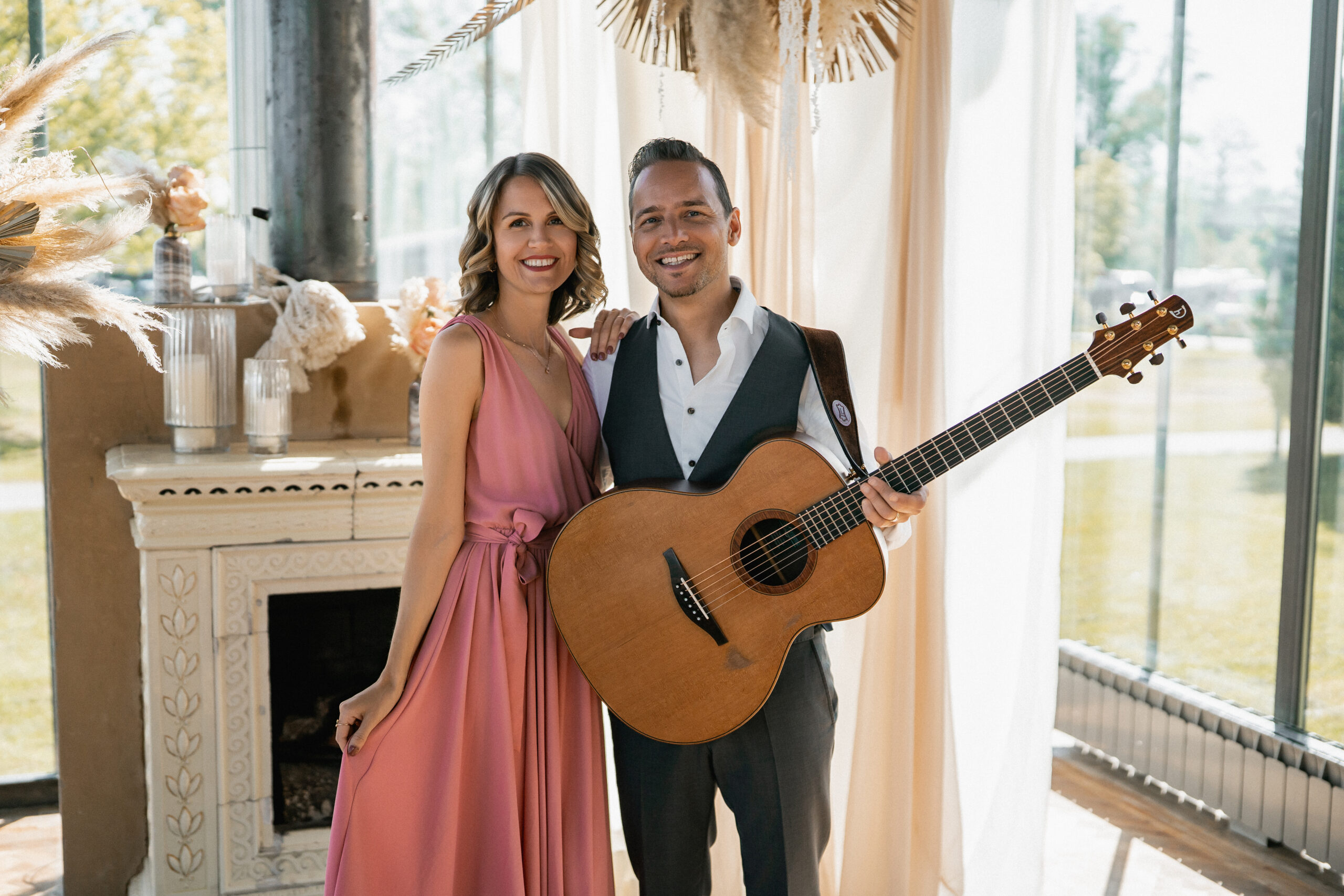 Eine lächelnde Frau in einem rosa Kleid steht neben einem Mann mit einer Akustikgitarre an einem Fenster - die perfekte Inspiration für Hochzeitsmusiker oder Hochzeitssänger in Süddeutschland.