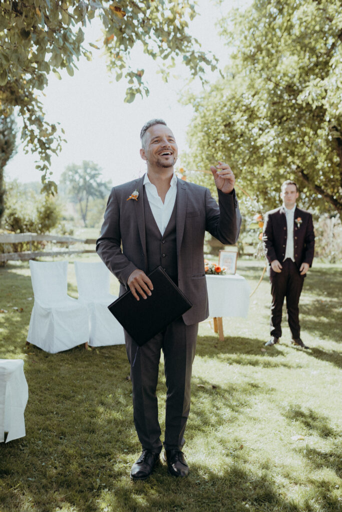 Ein Mann im Anzug steht im Freien in Südtirol, lächelt und hält eine Mappe in der Hand - perfekt gekleidet als Hochzeitssänger oder Trauredner, während im Hintergrund ein weiterer Mann und Stühle zu sehen sind.