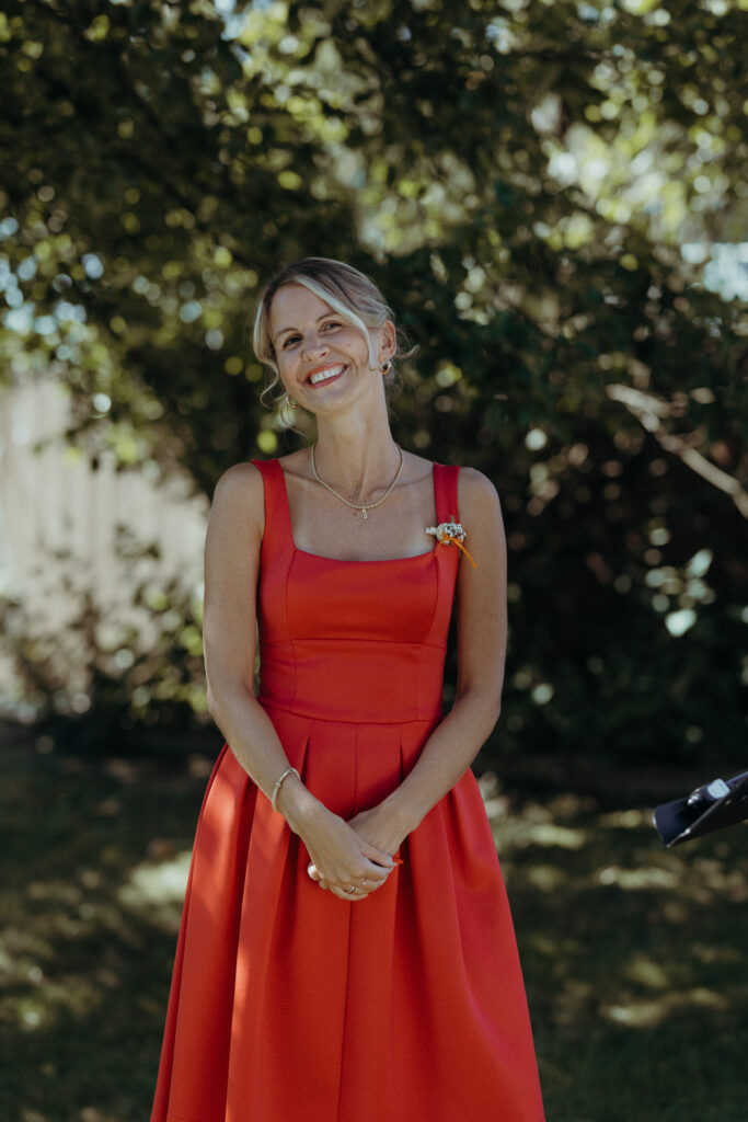 Eine lächelnde Frau in einem roten Kleid steht im Freien im Sonnenlicht, mit Bäumen und Grün im Hintergrund - die perfekte Inspiration für Hochzeitsmusiker oder Trauredner in Süddeutschland.