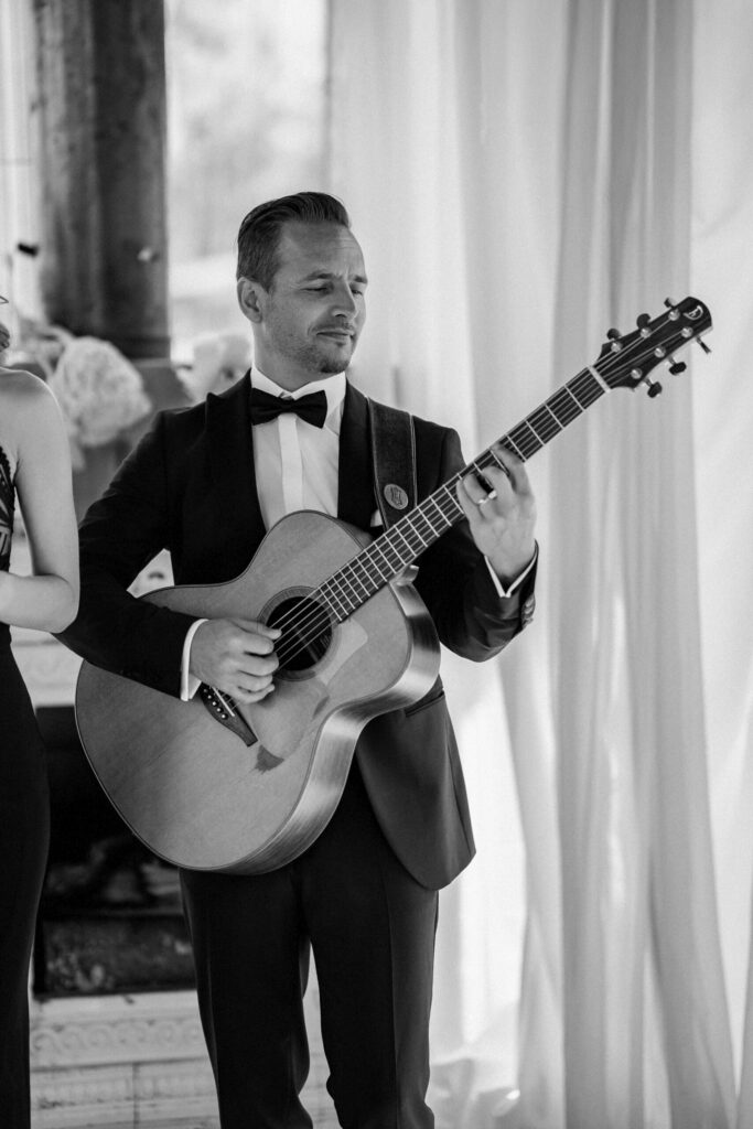 Ein Mann im Smoking spielt drinnen vor weißen Vorhängen auf einer Akustikgitarre – genau das Richtige, um jeder Freien Trauung eine besondere Note zu verleihen.