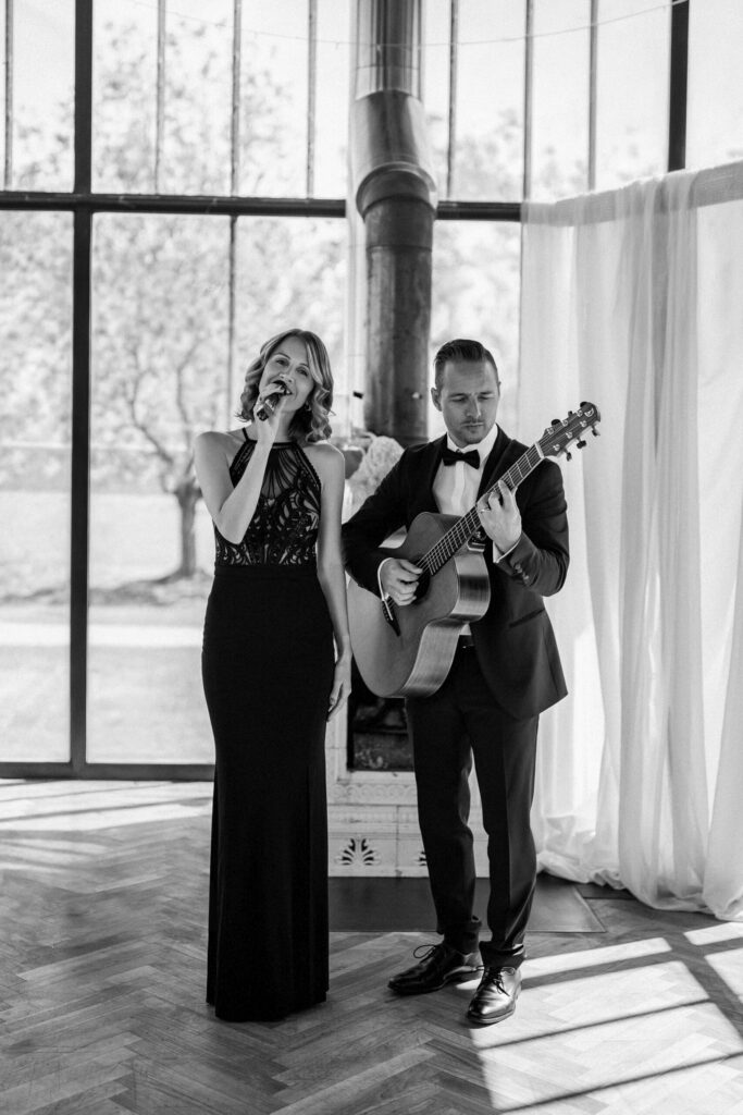 Eine Frau singt und ein Mann spielt Gitarre in festlicher Kleidung; sie treten in einem Raum vor großen Fenstern auf – eine perfekte Kulisse für eine freie Trauung mit Live-Musik.