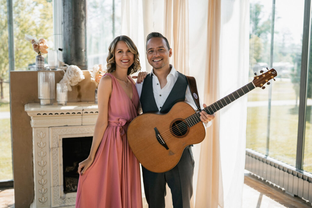 Eine Frau in einem rosa Kleid steht neben einem Mann in einer Weste, der eine Akustikgitarre in der Hand hält; beide lächeln in einem Raum – vielleicht feiern sie gerade mit ihrem freien Trauredner nach einer fröhlichen Zeremonie.