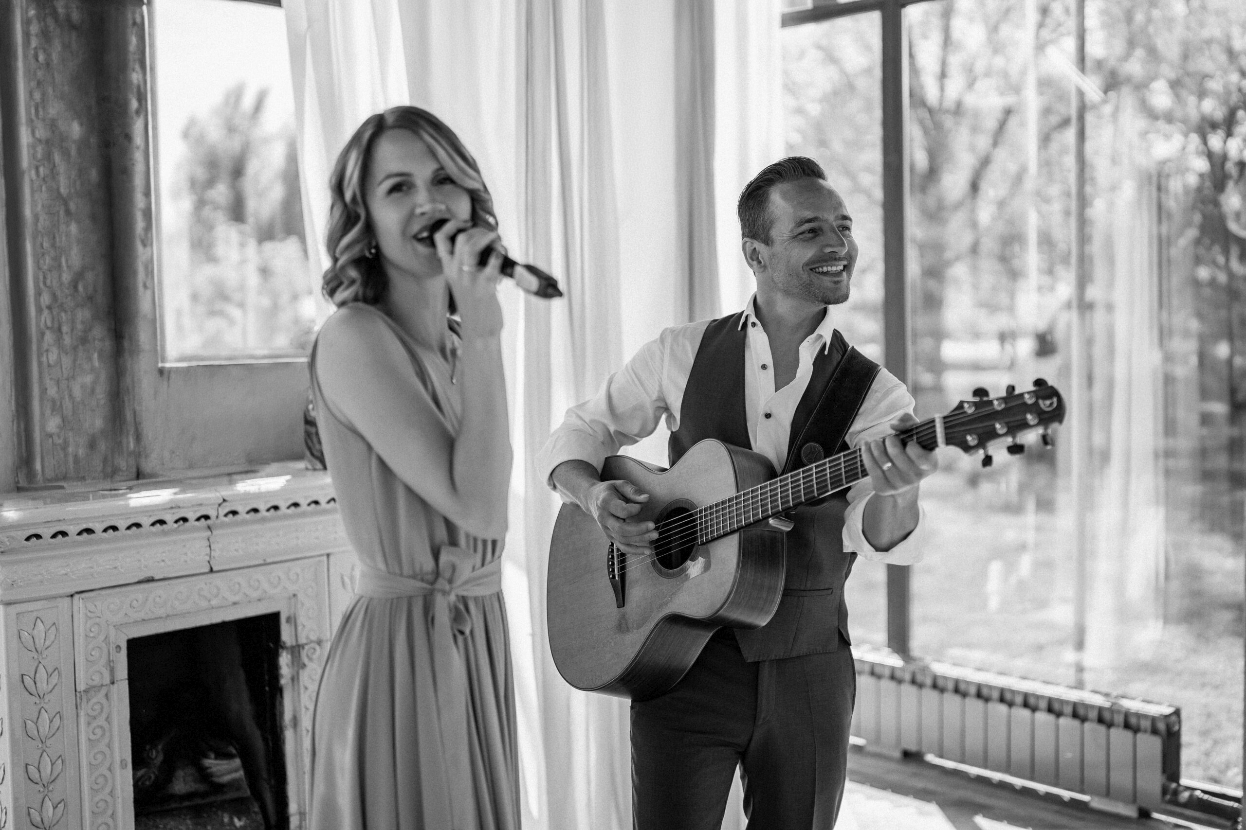 Eine Frau singt in ein Mikrofon, während ein Mann neben ihr auf einer Akustikgitarre spielt; beide lächeln.