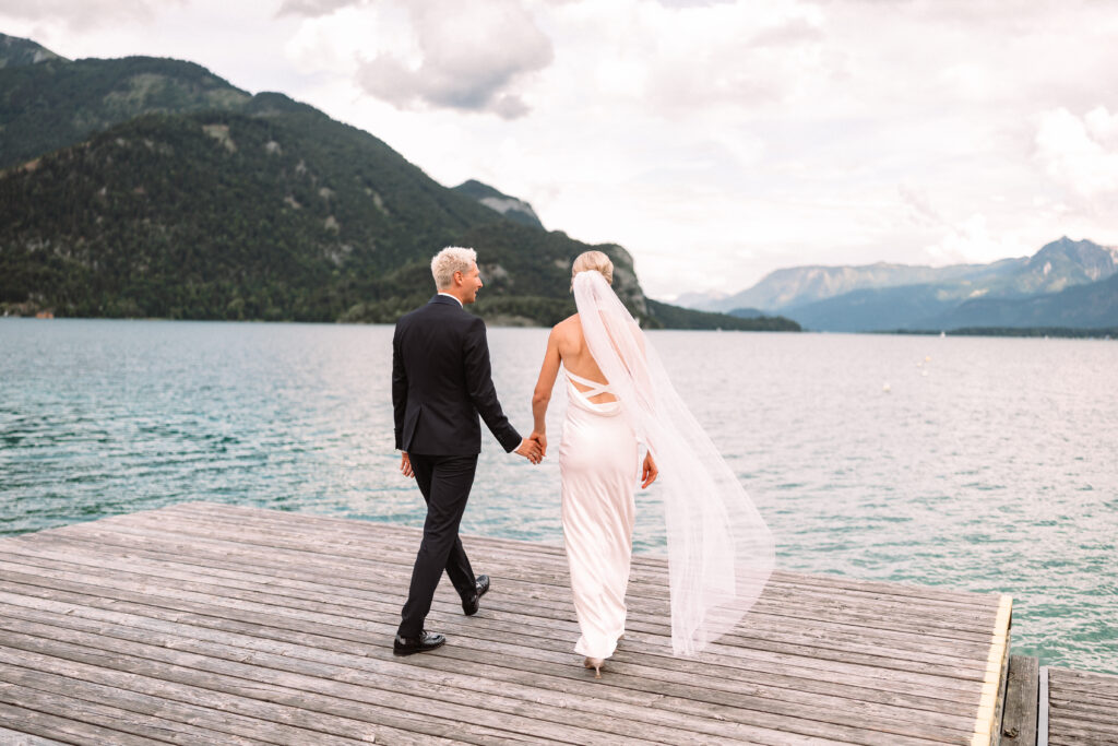 Ein Paar in Hochzeitskleidung spaziert Hand in Hand auf einem Steg an einem See entlang, im Hintergrund sind Berge zu sehen.