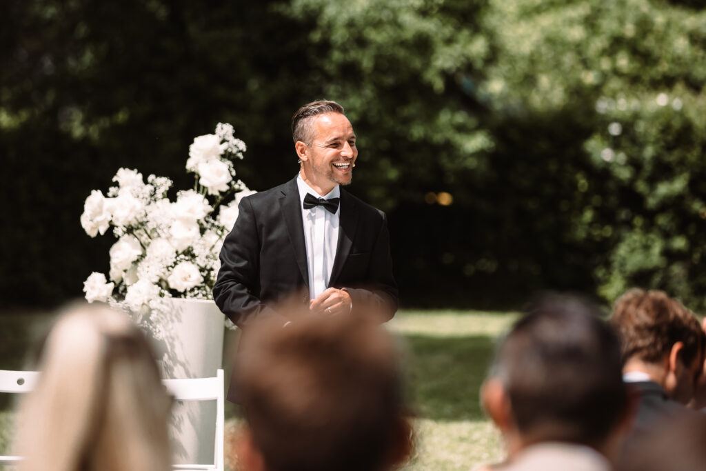 Ein Mann in einem schwarzen Smoking steht im Freien neben weißen Blumen und lächelt in Richtung eines sitzenden Publikums.